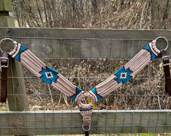 May include: A tan and turquoise braided horse breast collar with leather straps and metal rings. The breast collar is hanging on a wooden fence.