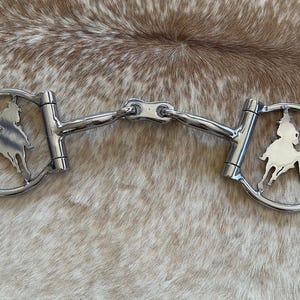 May include: A silver barrel racing bit with a cutout design of a horse and rider. The bit has a curved mouthpiece and two rings on either side.