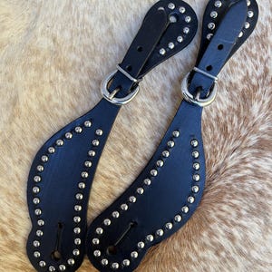 May include: A pair of black leather spur straps with silver studs. The straps have a buckle and a loop for attaching to a spur.