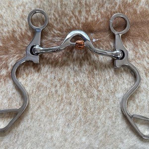 May include: A polished silver horse bit with a copper roller in the center. The bit has a curved design with a ring at each end and a curved, looped design. The background is a light brown and white speckled pattern.
