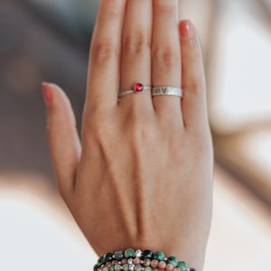 Peut inclure: Une main présentant des bijoux, dont une bague en argent avec une pierre rouge et un anneau en argent avec la lettre "A". Plusieurs bracelets de perles vertes, noires et oranges ornent le poignet. Les ongles sont peints d'un vernis rose clair.