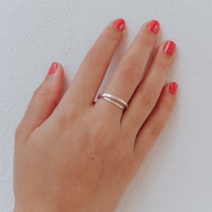 Könnte beinhalten: Ein silberner Doppelbandring mit gehammerter Textur. Der Ring wird an einer Hand mit rotem Nagellack vor weißem Hintergrund getragen. Der Schmuck ist schlicht und elegant.