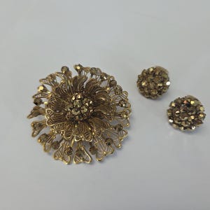 May include: A gold-toned vintage jewelry set featuring a large floral brooch and two matching round stud earrings. The brooch has an openwork design with a central cluster of stones. The earrings are also adorned with small stones.