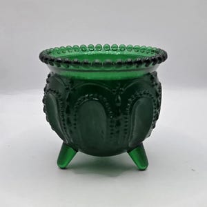 May include: A small, deep green glass bowl featuring a rounded body with three short legs. The exterior surface is decorated with a repeating embossed arch pattern and vertical beaded lines, while the rim is finished with a delicate beaded edge.