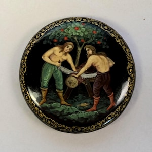 May include: A round black lacquer box featuring a hand-painted scene of two shirtless figures sawing a log under a fruit tree. The lid is decorated with a gold filigree border pattern. The figures wear green and brown trousers with boots.