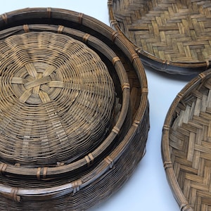May include: A set of three round, dark brown woven bamboo baskets. One basket is deep with a circular base pattern, while the other two are shallow, flat trays featuring a tight, intricate herringbone weave pattern throughout their surfaces.
