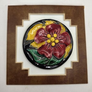 May include: A circular ceramic tile featuring a raised relief of a deep red flower with yellow centers and green leaves against a black background, mounted within a square brown wooden frame with a stepped inner border design.