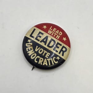 May include: A vintage political campaign button with a red and black color scheme. The button reads "LEADER VOTE DEMOCRATIC" in bold, white letters. The top section is red with the words "LEAD WITH" and stars.
