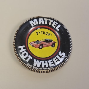 May include: A round metal pinback button featuring a black border with the white text MATTEL HOT WHEELS. The center has a yellow circle with a red illustration of a Python car. Text at the bottom reads 1967 Mattel Inc. Hong Kong.