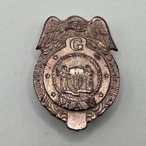 May include: Vintage metal badge with an eagle at the top and a circular emblem in the center. The emblem features two figures flanking a shield and the letter "G". The badge has a tarnished, antique appearance.