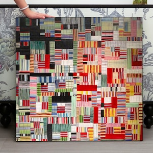 Puede incluir: Una pintura abstracta cuadrada con un marco negro. La obra de arte presenta un diseño de patchwork de formas rectangulares coloridas en rojo, amarillo, azul, verde y negro. La pintura está sostenida por una mano, con papel pintado floral en el fondo.