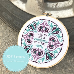 Peut inclure: Un motif de point de croix avec une citation du film "Fast and Furious" qui dit "Je vis ma vie à un quart de mile à la fois - Dominic Toretto". Le motif est encadré dans un cercle en bois et présente un motif floral dans des tons de violet, turquoise et vert.