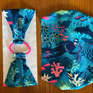 Puede incluir: Dos diademas de tela con fondo azul y una escena submarina colorida. Una diadema est&aacute; doblada y la otra est&aacute; plana. Ambas diademas tienen una banda el&aacute;stica rosa.