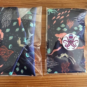 Puede incluir: Dos bolsas de tela con fondo azul y una escena submarina colorida. Las bolsas están empaquetadas en bolsas de plástico transparentes. La escena incluye corales, peces y un pulpo.