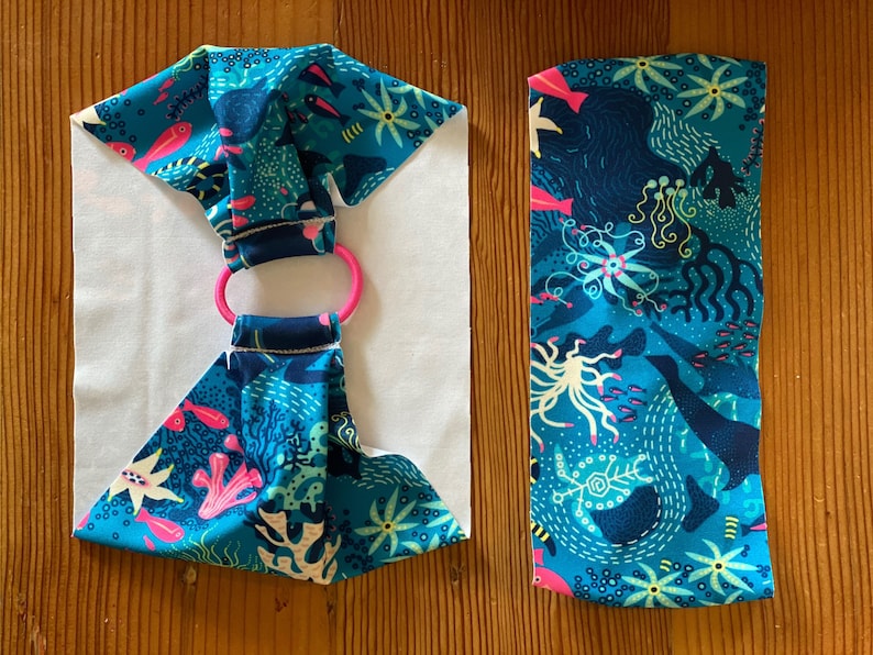 Puede incluir: Una diadema de tela azul y rosa con fondo blanco y un anillo rosa. La tela tiene un patr&oacute;n de peces, corales y otras criaturas marinas.