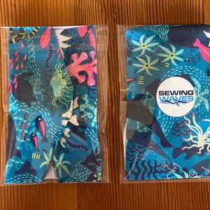Puede incluir: Dos diademas de tela empaquetadas con un fondo azul y una escena submarina colorida. Las diademas presentan peces rosas y azules, corales y algas. Una diadema tiene un c&iacute;rculo blanco con el texto "Sewing Waves".