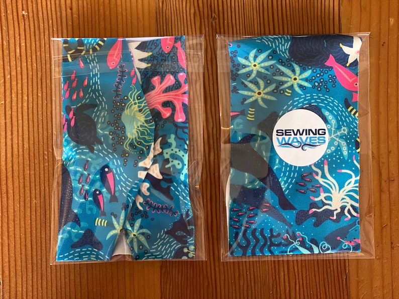 Puede incluir: Dos diademas de tela empaquetadas con un fondo azul y una escena submarina colorida. Las diademas presentan peces rosas y azules, corales y algas. Una diadema tiene un c&iacute;rculo blanco con el texto "Sewing Waves".