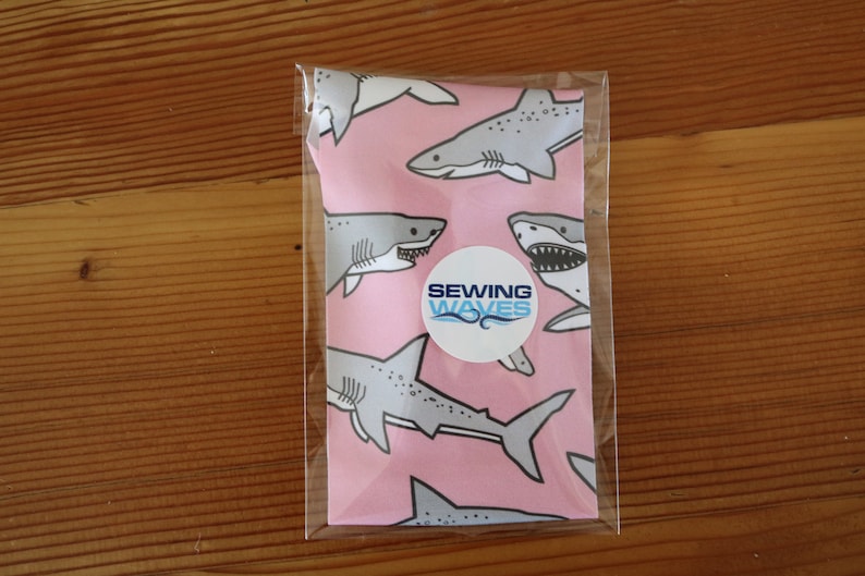 May include: A pink fabric with a white shark pattern and the text "Sewing Waves" on a white circle.