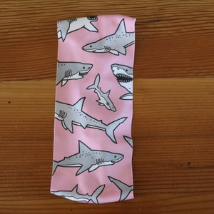 May include: Pink headband with a repeating pattern of gray sharks. The sharks are in various sizes and positions, some facing left and some facing right.