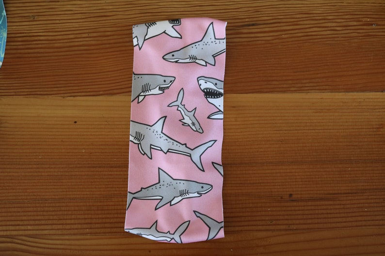May include: Pink headband with a repeating pattern of gray sharks. The sharks are in various sizes and positions, some facing left and some facing right.