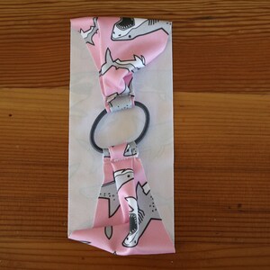 May include: Pink fabric headband with a white elastic band. The fabric has a pattern of gray sharks on a pink background.