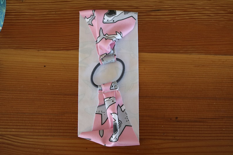 May include: Pink fabric headband with a white elastic band. The fabric has a pattern of gray sharks on a pink background.