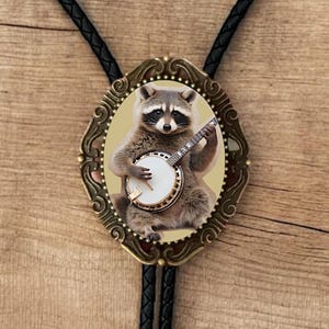 May include: A bolo tie featuring a brass-colored oval pendant with an image of a raccoon playing a banjo. The raccoon is brown and gray, and the banjo is white and gold. The tie has a black braided cord.