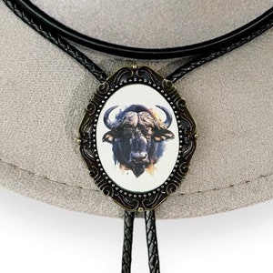 May include: A brown leather bolo tie with a silver-toned oval pendant featuring a black and white illustration of a buffalo head.
