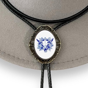 May include: A black bolo tie with a round, oval-shaped pendant featuring a white background with a blue floral design and a skull in the center. The pendant is surrounded by a gold-colored metal frame.