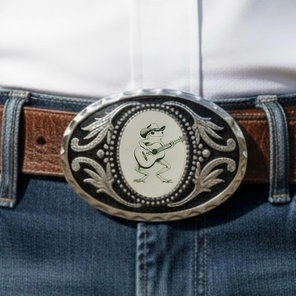 Puede incluir: Una hebilla de cinturón ovalada de color plateado con una ilustración central de una rana que lleva un sombrero de vaquero y toca una guitarra acústica. La hebilla tiene un fondo oscuro y texturizado con adornos plateados en forma de hojas.