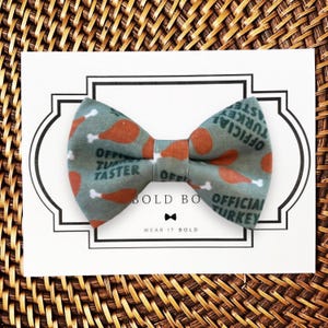 A dog bow tie with a pattern of turkeys and the text "OFFICIAL TURKEY TASTER" printed on it, placed on a white card with a black border.
