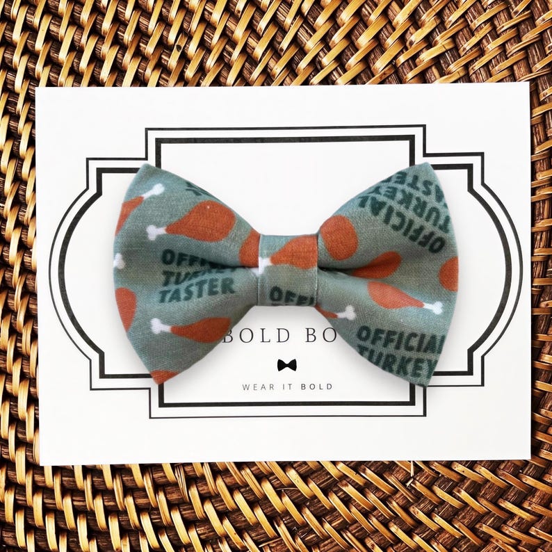 A dog bow tie with a pattern of turkeys and the text "OFFICIAL TURKEY TASTER" printed on it, placed on a white card with a black border.