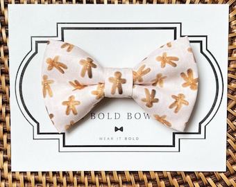 May include: A light pink fabric bow tie featuring a repeating pattern of metallic gold gingerbread men. The bow tie is attached to a white display card with the text THE BOLD BOW and WEAR IT BOLD printed in black.