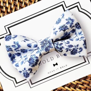 Op de afbeelding: Een witte vlinderdas met een blauw bloemenpatroon. De vlinderdas ligt op een witte kaart met de woorden "BOLD" en "WEAR IT BOLD". De vlinderdas is gemaakt van stof.