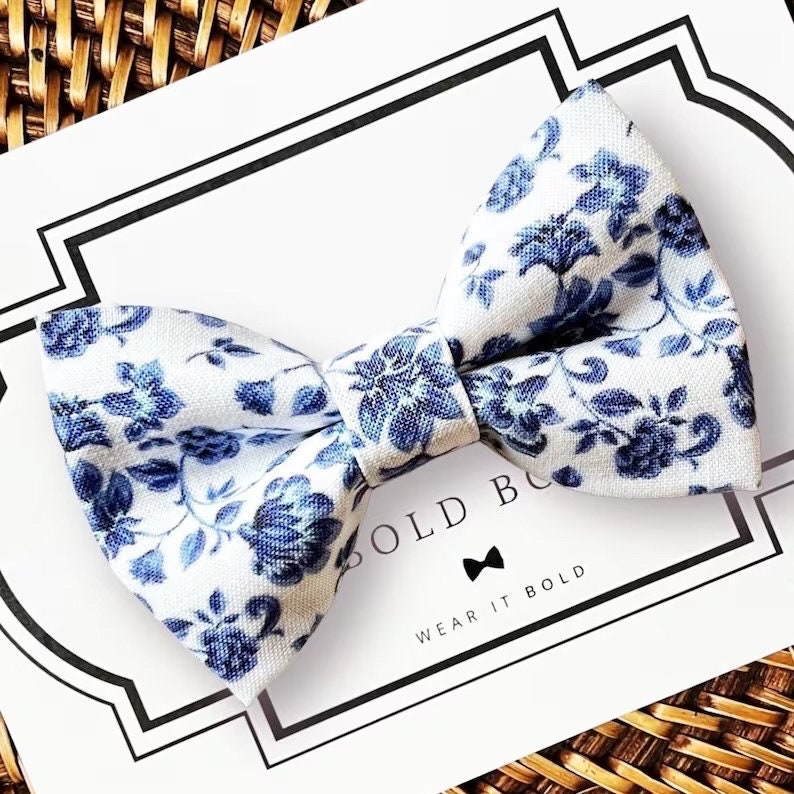 Op de afbeelding: Een witte vlinderdas met een blauw bloemenpatroon. De vlinderdas ligt op een witte kaart met de woorden "BOLD" en "WEAR IT BOLD". De vlinderdas is gemaakt van stof.
