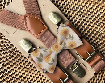 May include: A set consisting of brown elastic suspenders with tan faux leather accents and metal clips, paired with a matching bow tie featuring a cream-colored fabric printed with a repeating pattern of yellow sunflowers and green leaves.