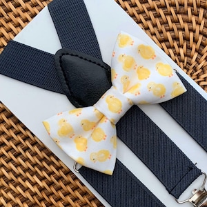 May include: A white bow tie featuring a repeating pattern of small yellow chicks, paired with dark navy blue elastic suspenders that have black leather-like tabs and silver-toned metal clips, all mounted on a white display card.