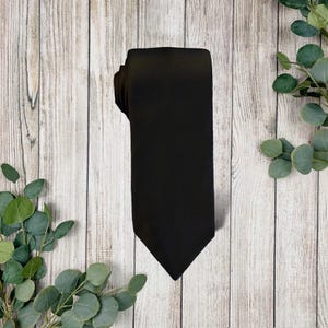 May include: A solid black necktie with a smooth, matte finish. The tie features a classic pointed tip at the bottom and a structured, rectangular shape that tapers slightly towards the top, displaying a clean and uniform dark fabric texture.