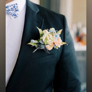 Op de afbeelding: Een donkergroene colbertjasje met een bloemenknop en een blauw-witte vlinderdas. De knop heeft perzik-, lichtblauwe en cr&egrave;mekleurige bloemen met groene bladeren. De tekst "@duetfloral" staat onderaan.