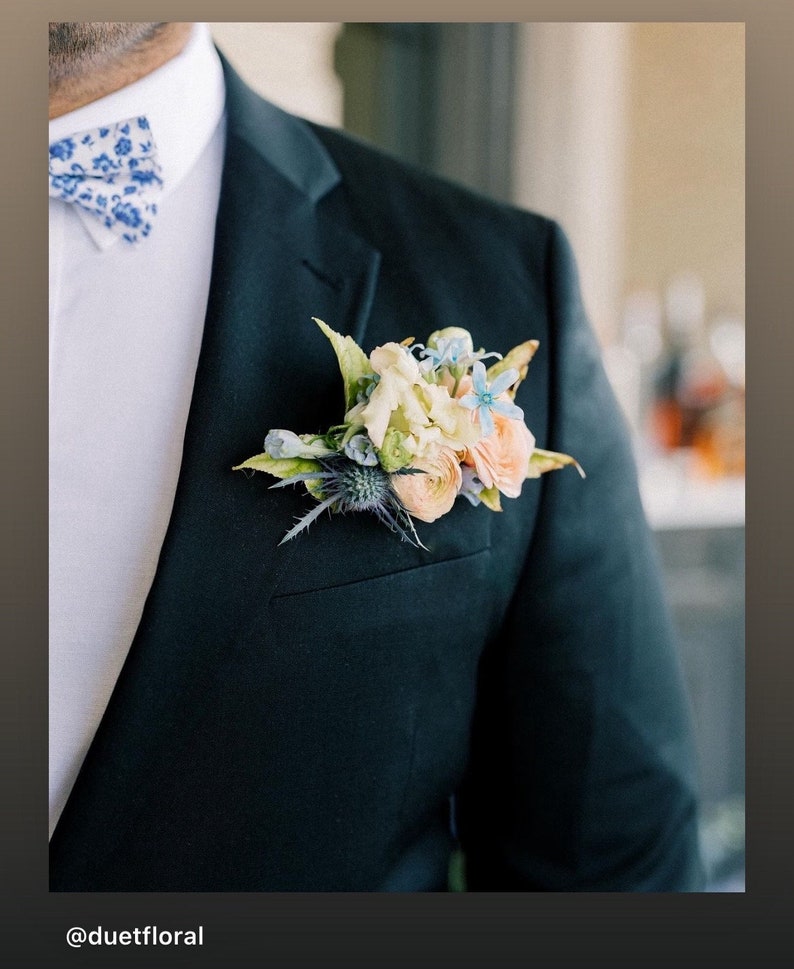 Op de afbeelding: Een donkergroene colbertjasje met een bloemenknop en een blauw-witte vlinderdas. De knop heeft perzik-, lichtblauwe en cr&egrave;mekleurige bloemen met groene bladeren. De tekst "@duetfloral" staat onderaan.
