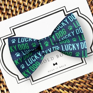 以下が含まれることがあります： ライトブルーとグリーンの「LUCKY DOG」という文字と小さな肉球のアイコンが繰り返されるパターンが特徴のネイビーブルーの蝶ネクタイ。蝶ネクタイは「BOLD BOW」および「WEAR IT BOLD」という文字が印刷された白い台紙の上に置かれています。