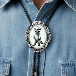May include: A bolo tie featuring an oval silver-toned metal frame with intricate scrollwork detailing. The center insert displays a black and white illustration of a raccoon wearing a cowboy hat against a light off-white background.