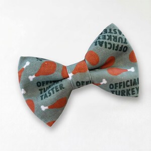 A Thanksgiving cat bow tie with a pattern of orange and white turkey images, along with the text "OFFICIAL TASTER" printed on it.