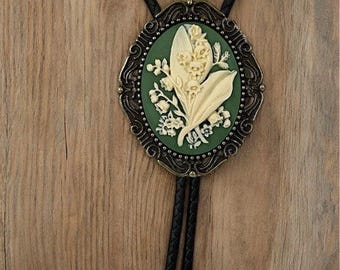 May include: A bolo tie featuring an oval-shaped pendant with an ornate bronze-toned metal frame. The center displays a raised cream-colored floral relief of lily of the valley flowers set against a solid dark green background. The cord is black braided leather with matching bronze-toned metal tips.