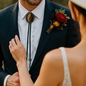 A dark navy suit with a white dress shirt, a bolo tie with a tiger's eye stone, and a red rose boutonniere. A woman's hand gently touches the man's chest, wearing a ring. The image captures a wedding or formal event.