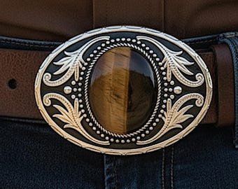 May include: An oval belt buckle featuring a central polished tiger's eye gemstone surrounded by a silver-toned metal frame with intricate engraved leaf patterns, beaded borders, and a dark recessed background, attached to a brown leather belt.