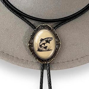 May include: A bolo tie featuring an oval-shaped centerpiece with a bronze-toned ornate metal frame. The center inlay displays a black and white illustration of a fish jumping out of water against a light beige background, attached to a black cord.