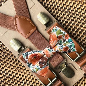 Könnte beinhalten: Ein Set aus braunen elastischen Hosenträgern mit beigen Kunstlederakzenten und goldfarbenen Metallclips, kombiniert mit einer passenden Fliege mit weißem Blumenmuster in Orange, Braun und Grün, befestigt auf einer rechteckigen Karte.