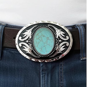 Puede incluir: Hebilla de cinturón ovalada con una piedra turquesa en el centro. La hebilla tiene un borde plateado con detalles negros y diseños de hojas decorativas. Se adjunta a un cinturón de cuero marrón y vaqueros azules.