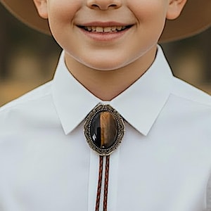 May include: A close-up of a bolo tie with a brown and gold oval stone set in an ornate, antique-style metal frame. The tie features a braided brown leather cord. The bolo tie is worn over a white collared shirt.